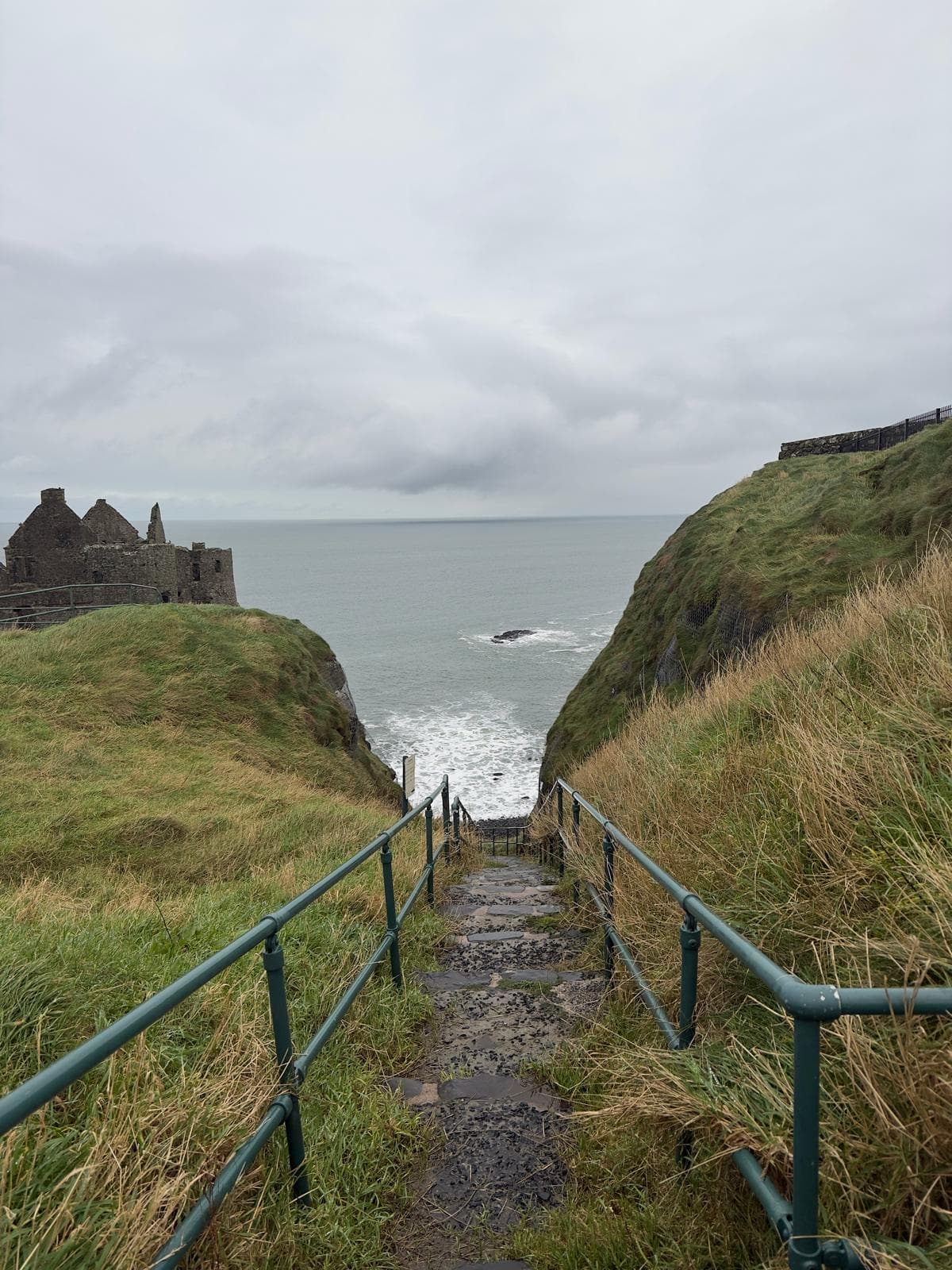 Railed steps down a grassy cliff to the sea, beside castle ruins